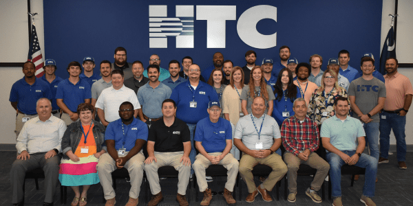 Group of HTC employees poses in front of wall with a large HTC logo and American and South Carolina flags on each side.
