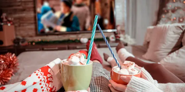 Two people with mugs of hot chocolate watch holiday movies in a cozy living room.